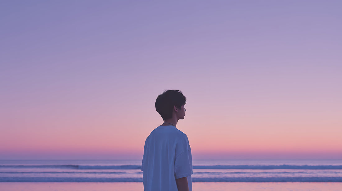 Ein junger Mann blickt bei Sonnenuntergang oder Sonnenaufgang über das ruhige Meer, die Szene ist in lila und rosa Tönen gehalten, was Ruhe und Reflexion ausstrahlt. Ideal zur Darstellung von Stressabbau und Achtsamkeit.