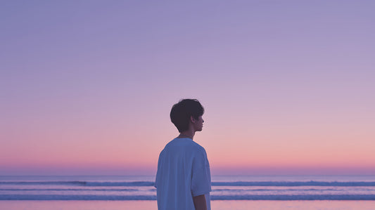 Ein junger Mann blickt bei Sonnenuntergang oder Sonnenaufgang über das ruhige Meer, die Szene ist in lila und rosa Tönen gehalten, was Ruhe und Reflexion ausstrahlt. Ideal zur Darstellung von Stressabbau und Achtsamkeit.