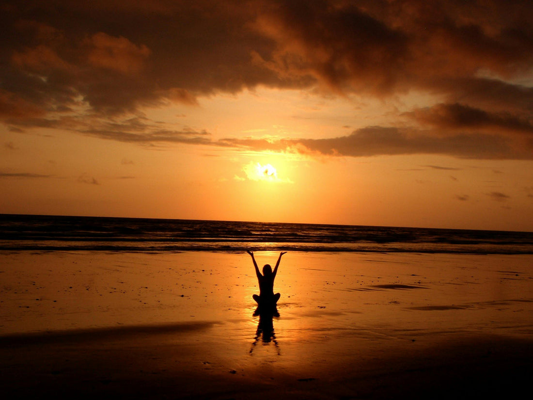 Person am Strand mit erhobenen Armen bei Sonnenuntergang über dem Meer.