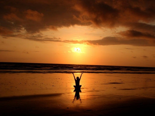 Person am Strand mit erhobenen Armen bei Sonnenuntergang über dem Meer.